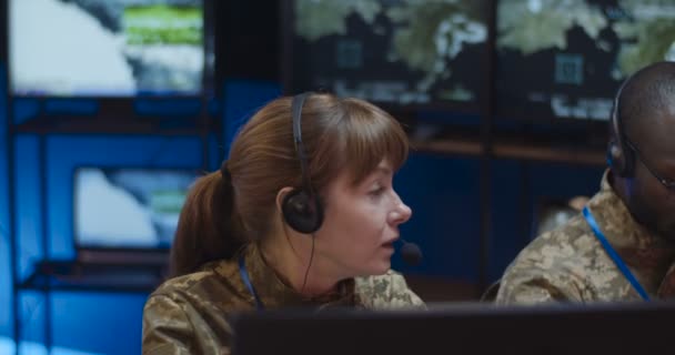 Homem e mulher multiétnicos, militares despachantes colegas em fones de ouvido sentados na sala de monitoramento com telas e controlando a operação das tropas. Dois oficiais de corridas mistas cooperando na sala de controle . — Vídeo de Stock