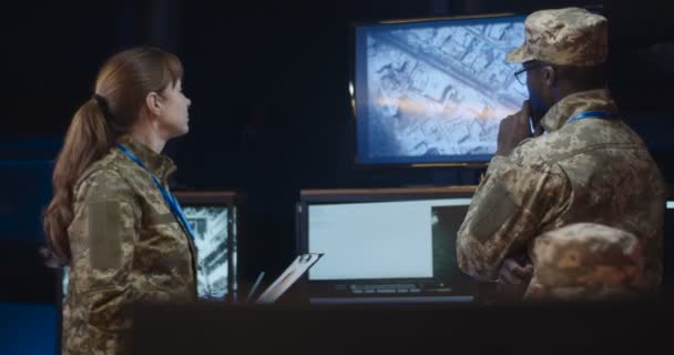 Afro-Amerikaanse man-generaal in camouflageuniform staat in de controlekamer en kijkt hoe troepen ontwrichten op schermen. Blanke vrouw komt en toont een aantal documenten met daa en info. — Stockvideo