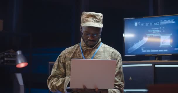 Portraitaufnahme eines afroamerikanischen, gut aussehenden jungen Armeegenerals, der im Überwachungsraum steht und Laptop-Computer benutzt. Soldat blickt geradeheraus und lächelt in die Kamera. — Stockvideo