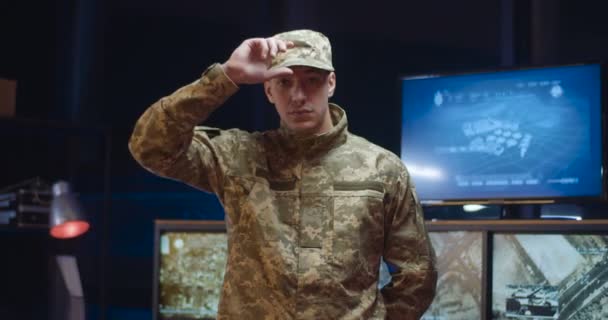 Retrato filmado de branco bonito jovem soldado em uniforme de camuflagem tirando a tampa e olhando para a câmera na sala de monitoramento. Homem militarista posando no escritório de controle . — Vídeo de Stock