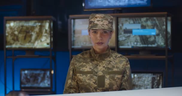 Güzel beyaz kadın askerin portresi kontrol odasında oturmuş kameraya neşeyle gülümsüyor. Kamuflaj üniformalı ve şapkalı güzel bir kadın asker olarak çalışıyor. Ordu konseptinde bir kız — Stok video