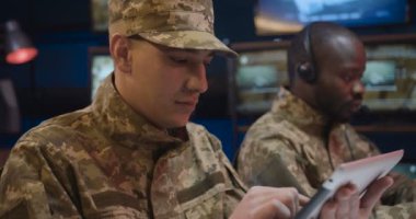 İzleme odasındaki tablet cihazında, üniformalı, şapkalı, kaydırmalı ve sörf yapan beyaz askerlere yaklaş. Afro-Amerikalı erkek meslektaşım arka plan üzerinde çalışıyor..