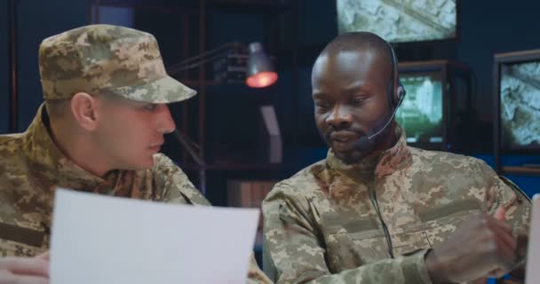 Multiethnische männliche Mitarbeiter sitzen im Kontrollraum und studieren offizielle Dokumente mit geheimen Daten. Afrikanischer junger Fahrdienstleiter arbeitet und spricht mit kaukasischen Kollegen. — Stockvideo