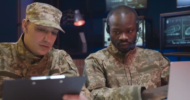 Gemengde-rassen militaire mannelijke werknemers zitten in het controlecentrum en het bestuderen van gegevens uit conflictgebieden. Afro-Amerikaanse man in headset typen op laptop computer terwijl Kaukasische soldaat lezen document — Stockvideo