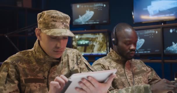 Kulaklıklı Afro-Amerikalı genç adam dizüstü bilgisayarda daktilo kullanıyor ve beyaz asker kontrol odasında tablet cihazını tıklatıyor. Üniformalı çok ırklı ordu subayları ofiste birlikte çalışıyorlar.. — Stok video
