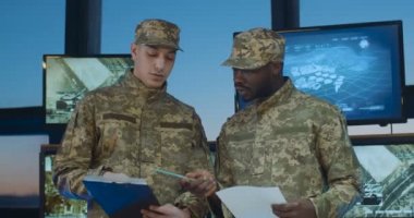 Üniformalı melez erkek askerler ofiste ekranlar ve bilgisayarlarla konuşuyor. Beyaz ve Afrikalı Amerikalı militanlar, veda ederken evrak okuyup el sıkışıyorlar..