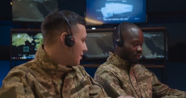 Peserta laki-laki ras campuran di tentara rekan kerja di headset duduk di ruang teknis militer dengan layar dan mengendalikan operasi perang. Dua orang ras campuran bekerja sama di ruang kontrol pasukan . — Stok Video