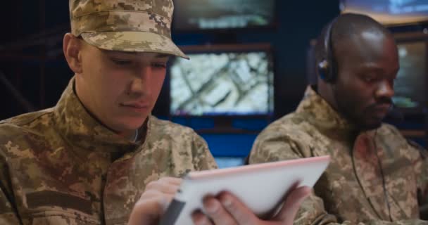 Gros plan du soldat masculin caucasien en uniforme et casquette tapant, défilant et surfant en ligne sur tablette dans le centre de contrôle. Afro-Américain collègue dans casque de travail sur fond . — Video