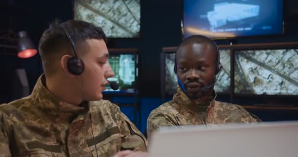 Despachadores militares masculinos multiétnicos colegas con auriculares sentados en la sala de monitoreo con pantallas y controlando la operación de guerra en la zona de conflicto. Dos hombres de razas mixtas que cooperan en la sala de control . — Vídeos de Stock