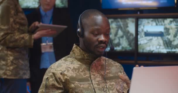 Афроамериканец в наушниках сидит за ноутбуком и разговаривает через видеочат в контрольном центре армии. Солдат говорит через веб-камеру в военном ведомстве. Офицер в диспетчерской . — стоковое видео