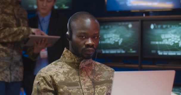 Portrét mladého afroamerického muže ve sluchátkách, který sedí u notebooku v monitorovací místnosti v armádě a pak se dívá do kamery. Detailní záběr mužského vojáka dispečera ve vojenském centru. — Stock video