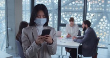 Ofiste akıllı telefon kullanarak koruyucu maske takan Asyalı genç bir iş kadınının portresi. Sağlık ve güvenlik hayatı kavramı, koronavirüs, virüs koruması, salgın hastalık.