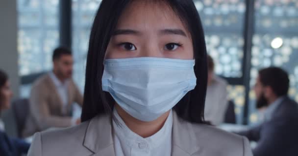 Close up face portrait of a young asian business woman wearing protective mask looking at camera in office. Concept of health and safety life, coronavirus, virus protection, pandemic. — Stock Video