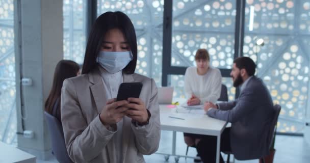 Portret van een jonge Aziatische zakenvrouw met beschermende masker typen, met behulp van smartphone op kantoor. Concept van gezondheid en veiligheid, coronavirus, virusbescherming, pandemie. — Stockvideo