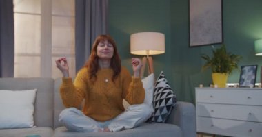 Kızıl saçlı güzel beyaz kadın loto masasında rahat oturma odasında koltukta oturmuş meditasyon yapıyor. Kadın evde yoga ve meditasyon yaparken dinleniyor ve dinleniyor..
