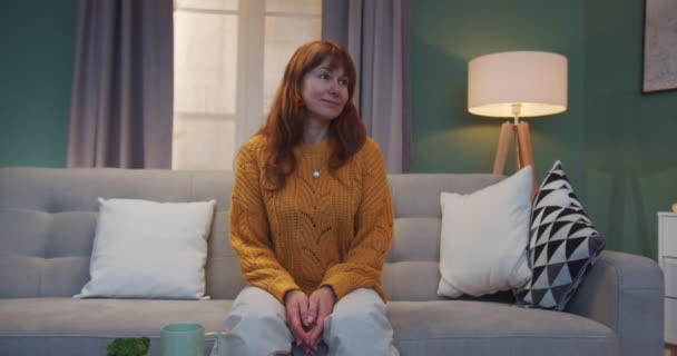 Портрет красивой белой рыжей женщины в желтом свитере, сидящей на диване в гостиной и улыбающейся в камеру. Женщина, отдыхающая дома на диване . — стоковое видео