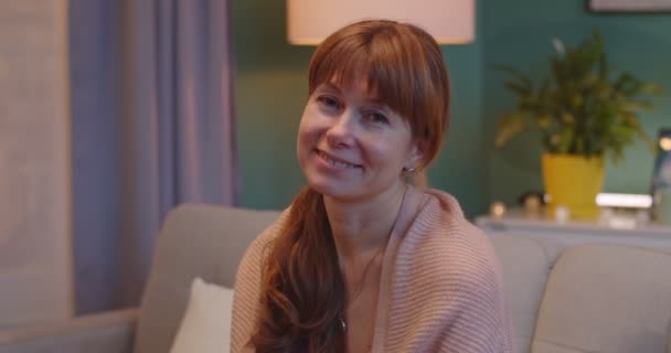 Primer plano de la bella mujer caucásica de mediana edad pelirroja sentada en el sofá en la acogedora sala de estar y sonriendo a la cámara sinceramente. Hermosa mujer hogareña mirando con sonrisa feliz en casa . — Vídeos de Stock