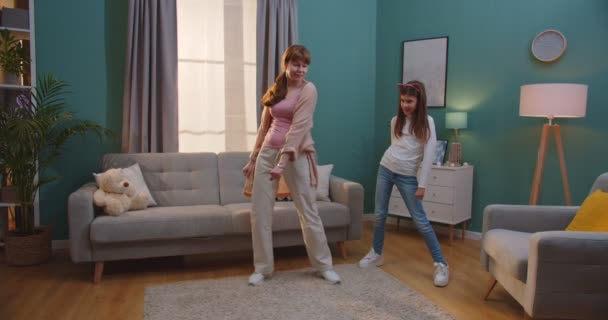 Veselá hezká běloška matka ukazuje taneční pohyby k roztomilé malé dceři, zatímco oni se baví spolu v obývacím pokoji. Žena a mladá dívka tančí o víkendu doma. Koncept mateřství. — Stock video