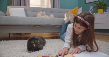 Komik kulakları olan beyaz tatlı bir kız yerde uzanıyor ve bebek kitabının sayfalarını çeviriyor ve yanında kedi oturuyor. Küçük bir çocuk kitap okuyor ve kedisiyle oturma odasında resim çekiyor..