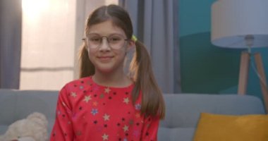 Evdeki kanepede otururken kameraya neşeyle gülümseyen, gözlüklü, beyaz, şirin genç kız portresi. Oturma odasında mutlu bir çocuk.