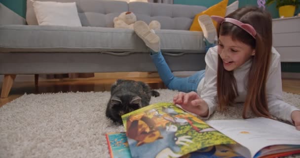 Kaukasisch klein schattig tienermeisje spelen met kittycat terwijl liggend op de vloer in de woonkamer en het lezen van boek voor kinderen. Mooi kind met kleurrijk sprookje en poesje rust thuis. — Stockvideo