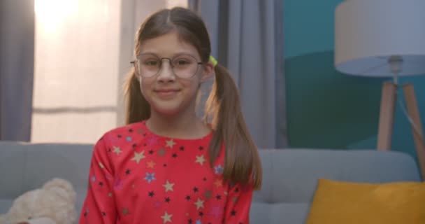 Portrait de petite adolescente blanche mignonne dans des lunettes souriant joyeusement à la caméra tout en étant assis sur le canapé à la maison. Enfant heureux dans le salon confortable . — Video