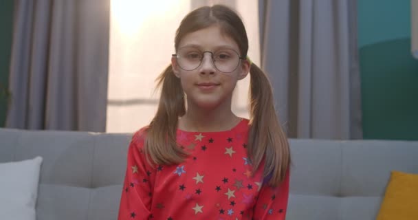 Portré felvétel kaukázusi kis aranyos tini lány szemüvegben fordult arc és vidáman mosolyog a kamera, miközben ül a kanapén otthon. Boldog kisgyerek a kényelmes nappaliban.. — Stock videók