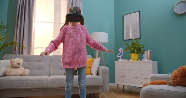 Mooi blank meisje met VR-brilletjes die in de woonkamer speelt in virtual reality. Klein grappig kind met een headset terwijl hij thuis met zijn handen zwaait. Wereld van virtualiteit concept. — Stockvideo
