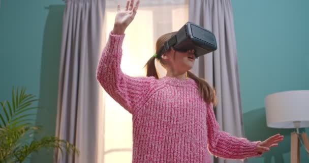 Kleine kaukasische niedliche und hübsche Mädchen mit VR-Brille stehen im Wohnzimmer und spielen in virtueller Realität wie etwas zu berühren. Kleines lustiges Kind mit Headset zu Hause. Konzept der Virtualität. — Stockvideo