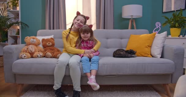 Hübsches kaukasisches Teenie-Mädchen umarmt kleine süße Schwester, während Schwestern auf der Couch im schönen Wohnzimmer sitzen. Zwei fröhliche und lächelnde Mädchen, die sich auf dem Sofa im Haus umarmen. Kind zu Hause. — Stockvideo