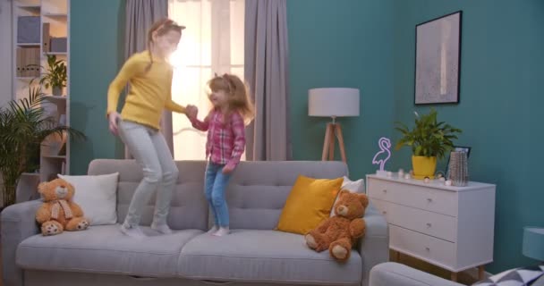 Vreugdevolle kleine schattige blanke meisjes in grappige oren springen op de bank in gezellige woonkamer. Mooie zusjes die plezier hebben, thuis spelen en lachen. Gelukkige jeugd. — Stockvideo