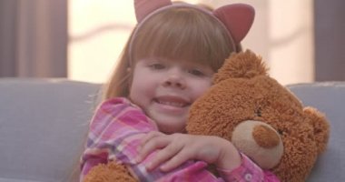 Komik kulaklı, sevimli, beyaz bir kızın, dar oyuncaklara sarıldığı ve koltukta otururken gülümsediği bir sahne. Evdeki oturma odasında oyuncak ayıyı kucaklayan güzel bir çocuğun portresi..