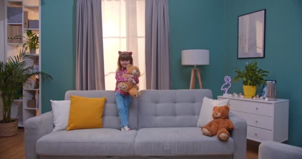 Portret małej białej dziewczyny w śmiesznych uszach stojącej na kanapie i trzymającej w domu zabawkę pluszowego misia. Mały dzieciak uśmiecha się na kanapie w salonie. — Wideo stockowe