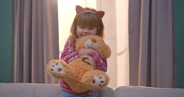 Portrait de jolie petite fille caucasienne aux oreilles drôles regardant la caméra tenant jouet d'ours en peluche à la maison. Petit enfant souriant sur le canapé dans le salon . — Video