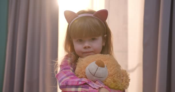 Nahaufnahme eines hübschen kleinen kaukasischen Mädchens mit lustigen Ohren, das direkt in die Kamera schaut, Teddybär zu Hause hält und umarmt. Porträt eines kleinen Kindes, das Spielzeug im gemütlichen Wohnzimmer umarmt. — Stockvideo
