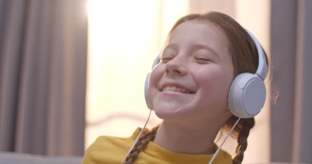 Close up de bonito pequena menina caucasiana em fones de ouvido ouvindo música e rindo enquanto sentado no sofá interior. Retrato de criança bonita desfrutando de música favorita na sala de estar em casa . — Vídeo de Stock