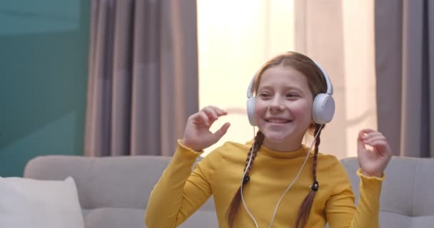 Linda chica caucásica pequeña en auriculares escuchando música y riendo mientras se sienta en el sofá interior. Primer plano de niña bonita bailando y divirtiéndose en la sala de estar en casa . — Vídeo de stock