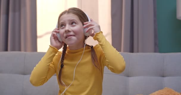 En söt liten flicka som tar på sig hörlurar och lyssnar på musik med leende. Barn i gul tröja njuter av favoritsång och leende medan du sitter på soffan hemma. — Stockvideo