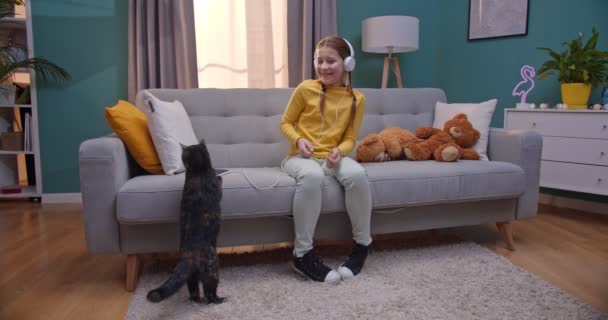 Caucasian small teen pretty girl in headphones listening to music and sitting on couch in living room. Cat jumping on sofa and kid caressing it at home. — Stock Video