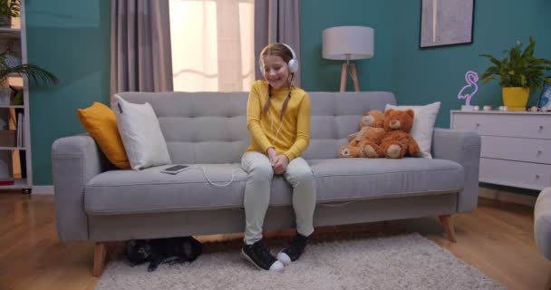 Καυκάσιος μικρός έφηβος όμορφο κορίτσι με ακουστικά ακούγοντας μουσική και κάθεται στον καναπέ στο άνετο σαλόνι. Γάτα κοιμάται κάτω από τον καναπέ στο σπίτι. — Αρχείο Βίντεο