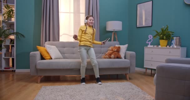 Kavkazský dospívající malá hezká dívka ve sluchátkách poslech hudby a tanec v útulném obývacím pokoji. Veselé roztomilé dítě tanec a baví se doma. — Stock video
