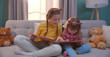 Komik kulaklı beyaz çocuklar kanepede oturur ve rahat oturma odasında kitap okurlar. Güzel küçük kız kardeşler masal okurlar. Evde büyüteçle ders kitabını araştıran kızlar.