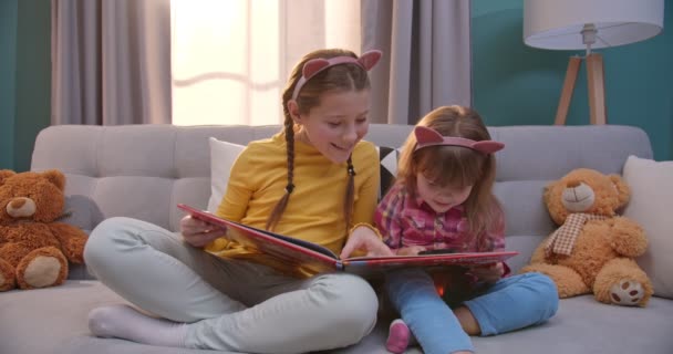 Petits enfants mignons caucasiens dans des oreilles drôles assis sur le canapé et le livre de lecture dans le salon confortable. De jolies petites sœurs lisent des contes de fées. Les filles explorant manuel avec loupe à la maison . — Video
