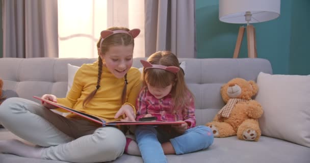 Кавказские милые маленькие дети в смешных ушах сидят на диване и читают книгу в гостиной. Красивые сестрички читают сказки и веселятся. Девушки изучают учебник с лупой дома — стоковое видео