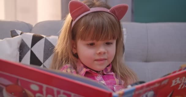 Detailní záběr kavkazské roztomilé malé dítě v legrační uši sedí na pohovce a čtení knihy v obývacím pokoji. Hezké dítě čte pohádky a usmívá se. Dívka zkoumání učebnice s lupou doma. — Stock video