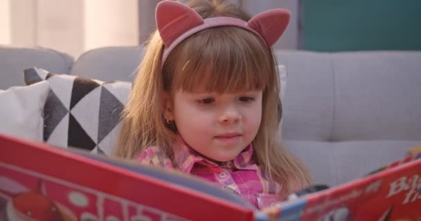 Close up de caucasiano bonito criança pequena em ouvidos engraçados sentado no sofá e ler livro na sala de estar. Um miúdo bonito lê contos de fadas e sorri. Menina explorando livro didático com lupa em casa . — Vídeo de Stock