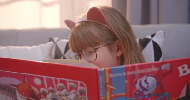 Close up of Caucasian cute little child in funny ears and glasses sitting on sofa and reading book. Pretty small girl flipping pages with smile. Kid read story at home. — Stock Video