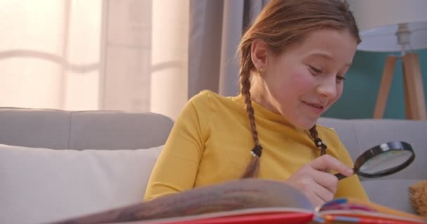 Caucasiano bonito adolescente criança rindo enquanto sentado no sofá e olhando para a câmera através de lupa. Um miúdo bonito a ler livros de contos de fadas e a sorrir. Menina sorrindo em casa. Leitor pequeno . — Vídeo de Stock