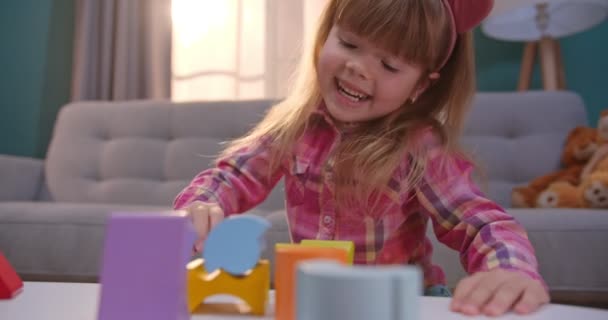 Caucásico pequeño niño bonito y lindo sentado en acogedora habitación agradable en la mesa y jugando con juguetes para bebés. Chica alegre divertirse y jugar en interiores. Niño pasando tiempo solo en casa . — Vídeo de stock