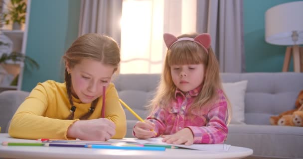 Kaukasiska små söta och söta systrar sitter i mysiga trevligt rum vid skrivbordet och färg bild med färgglada pennor. Glada tjejer som har roligt och leker inomhus. Barn som är ensamma hemma. — Stockvideo